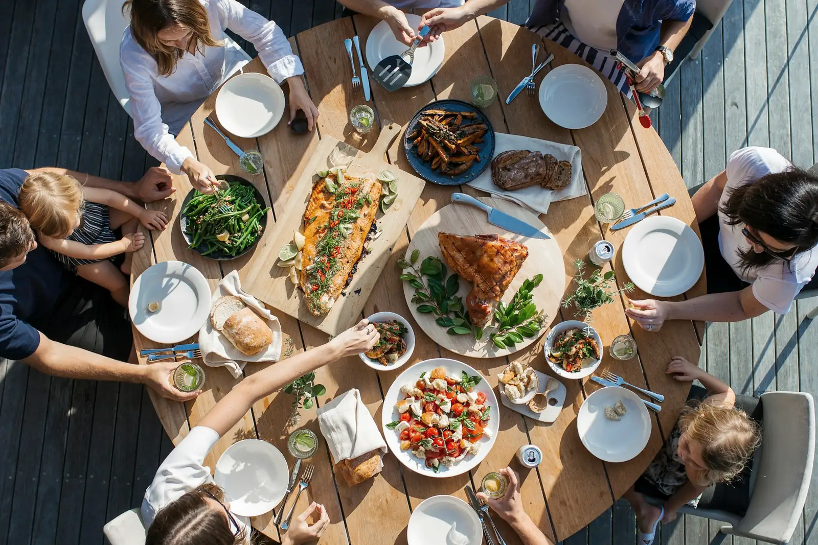 Seen from above, a round wooden table outdoors laid with shared plates of salmon, grilled vegetables, bread and salad, with adults and children reaching in to serve themselves